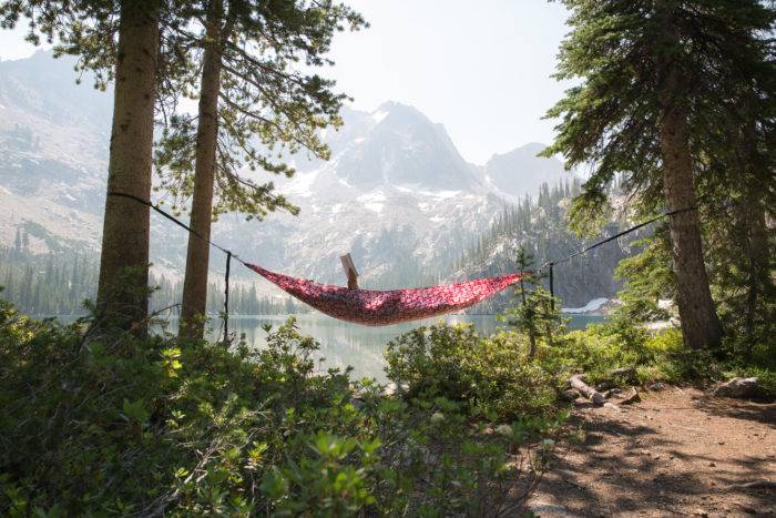 A person laying in red hammock in front of a lake with a mountain range behind. 
