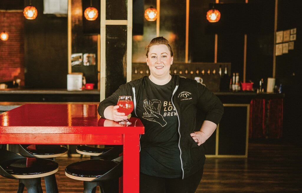 A woman standing in a brewery with her arm on a table holding a glass of red-hued beer.