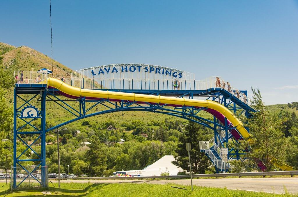 Water park visitors walk up stairs to access two waterslides near a sign that says Lava Hot Springs with a mountain in the background.