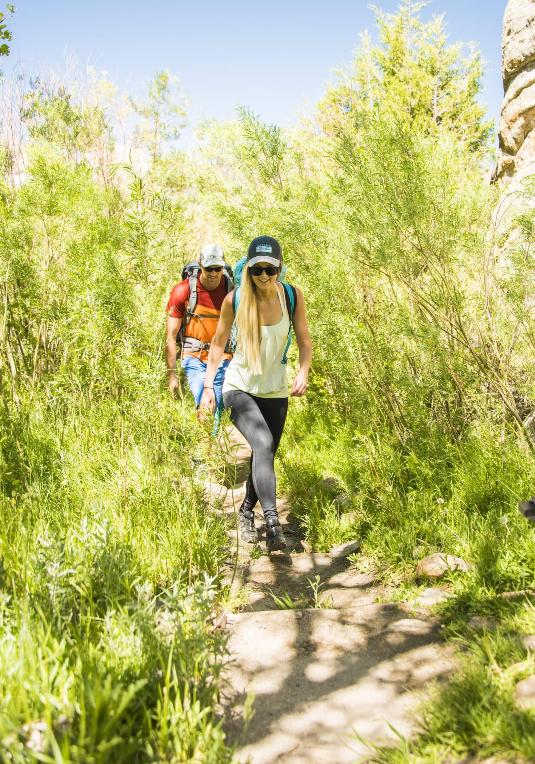 Two people hiking on a trail surrounded by tall shrubs and grass at City of Rocks National Reserve in the summer.