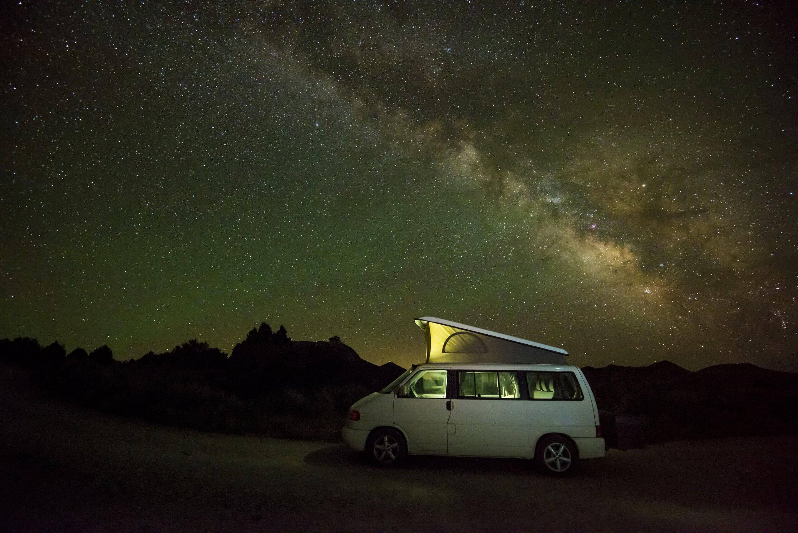 A camping van with a tent on the roof with the Milky Way seen in the starry night sky.