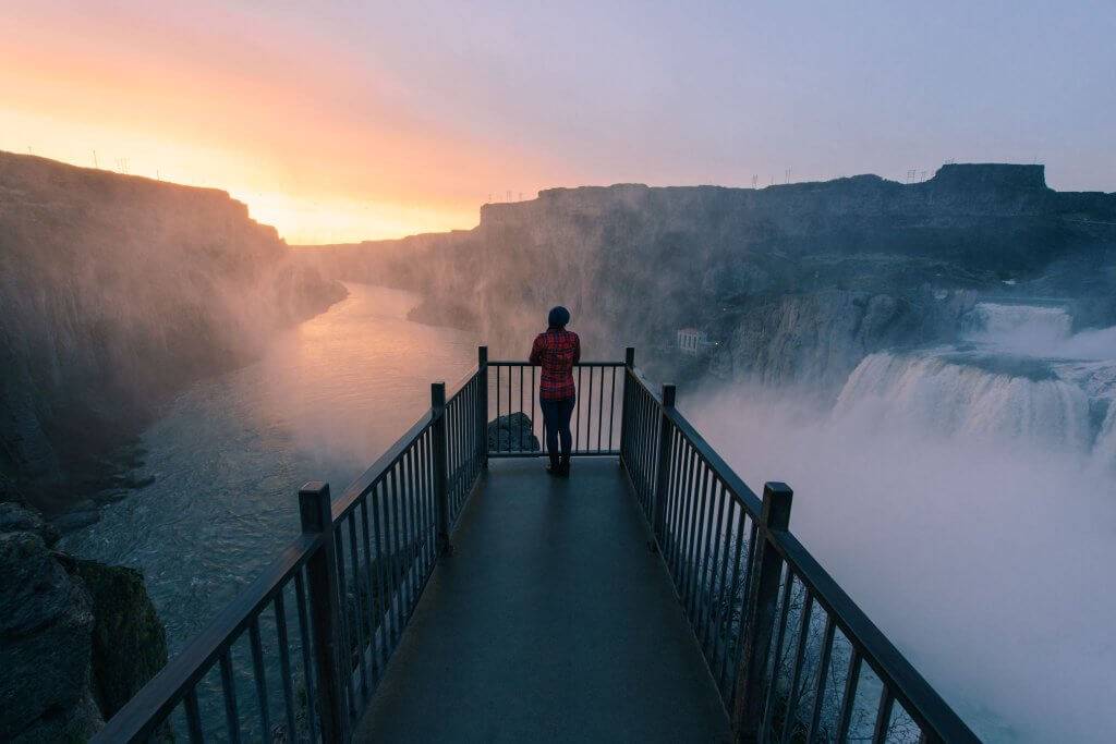 A woman stands at the end of a viewing deck, surrounded by mist from Shoshone Falls at sunset.