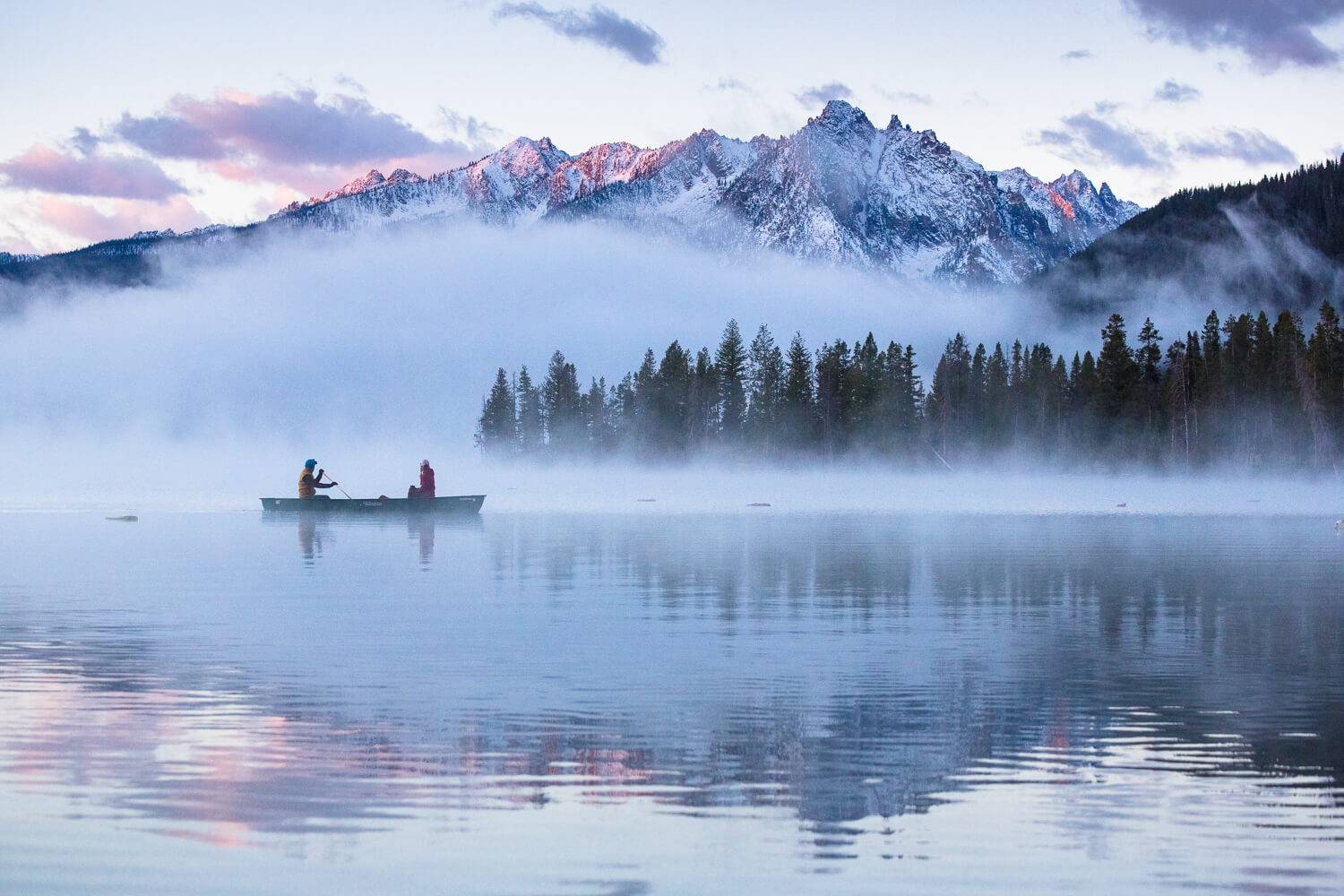two people sit in a boat on a lake surrounded by fog with snow-capped mountains in the background