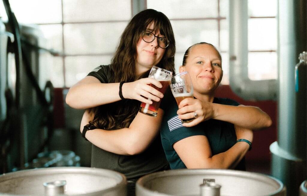 Two women clinking glasses with beer in them with beer brewing equipment surrounding them.