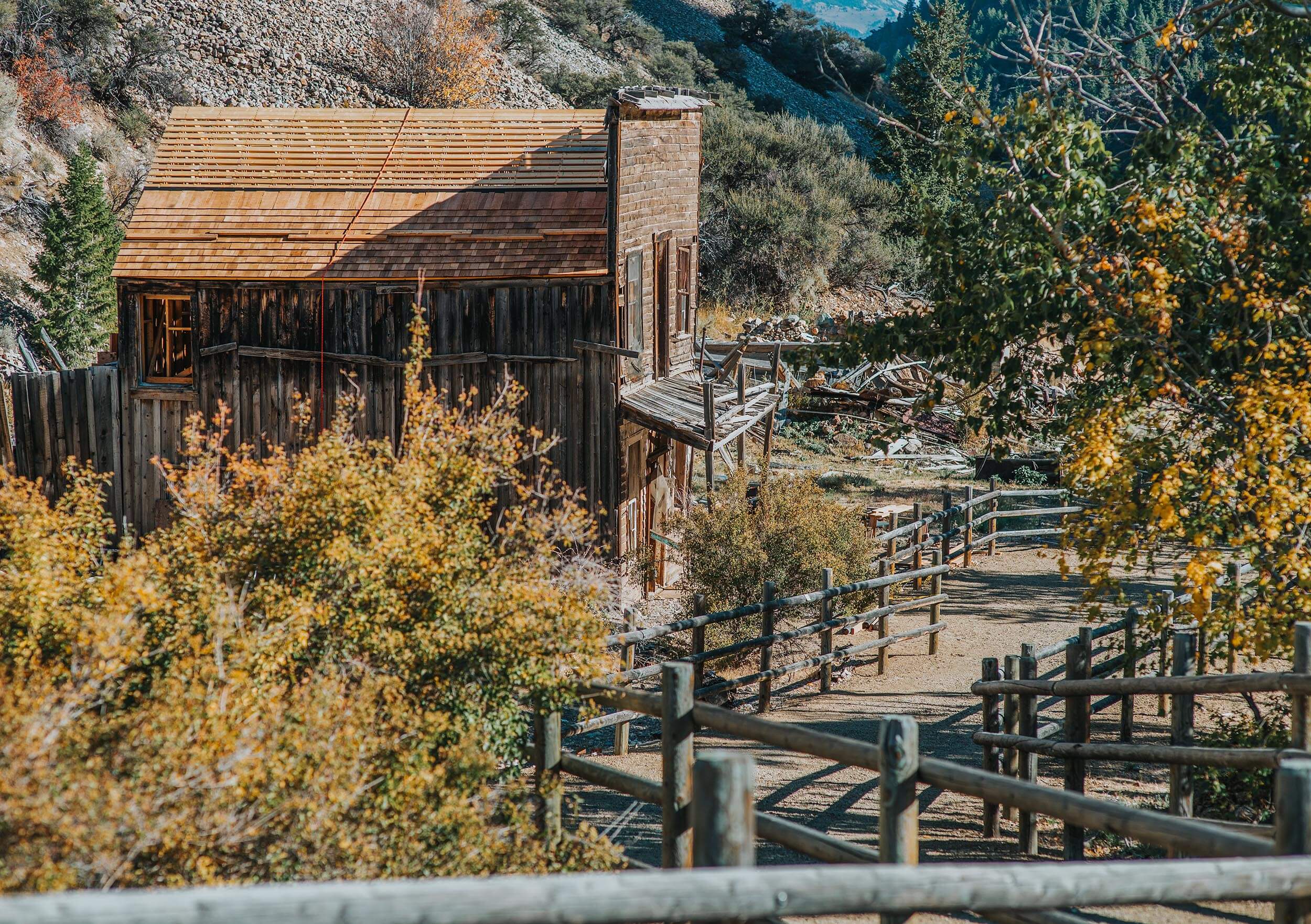 An old wooden and brick building beside a dirt path lined with fencing, surrounded by trees at Bayhorse Ghost Town.