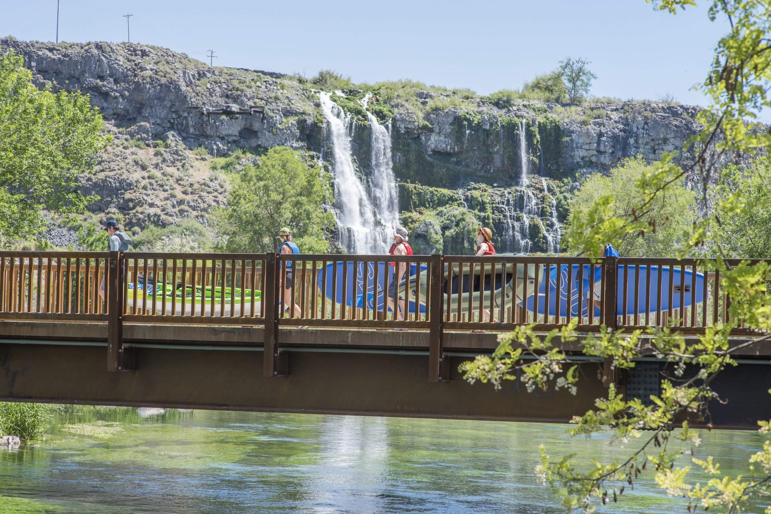 A group of people walking a cross a bridge carrying standup paddlenboards, and a waterfall in the background at Thousand Springs State Park.