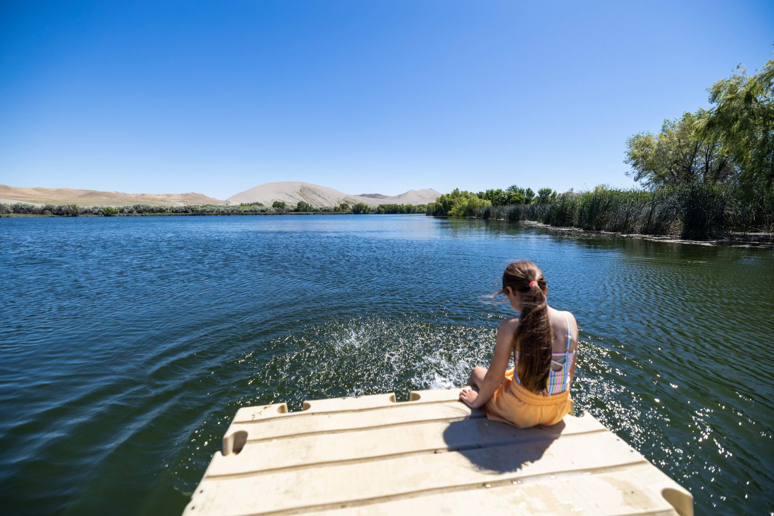 A girl sits on the edge of a dock with her feet in Dunes Lake at Bruneau Dunes State Park.