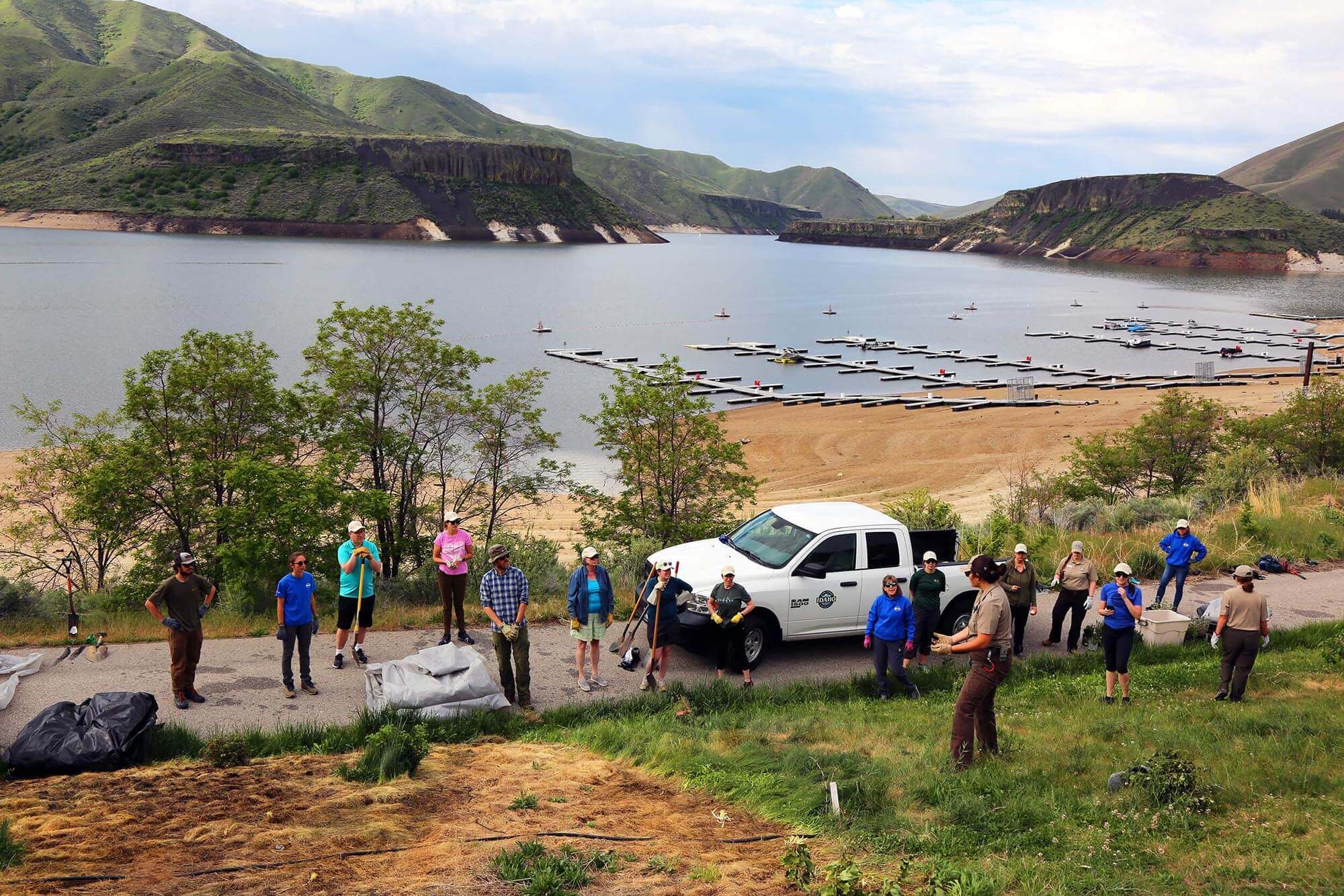 A group of volunteers holding gardening tools at a Travel With Care event and a beach, docks, body of water and mountains in the background.