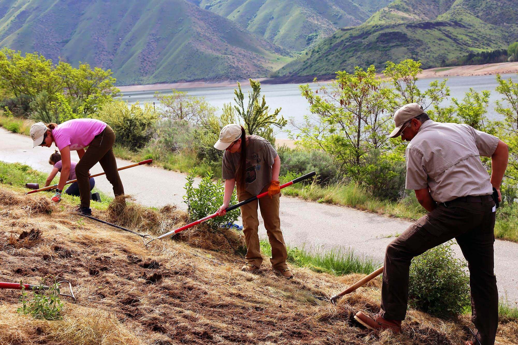 Three people at a Travel With Care event holding garden hoes standing on a sloped hill and a paved path, a body of water and mountains in the background.