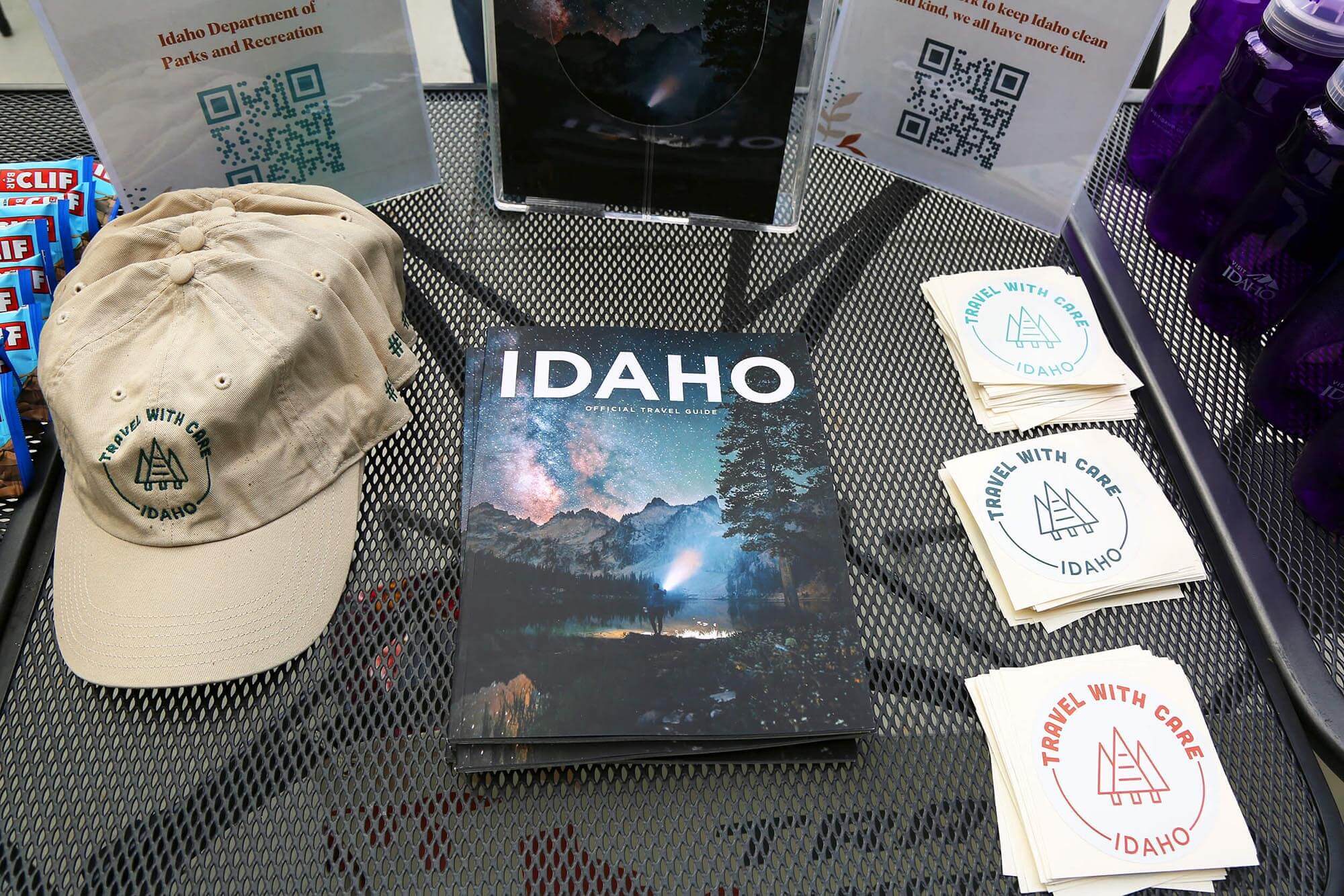 A metal table with Travel With Care hats, stickers and water bottles positioned around a stack of 2023 Official Idaho Travel Guides.