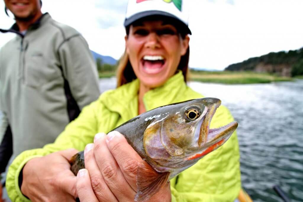 A woman in a neon green jacket smiles while holding a cutthroat trout.