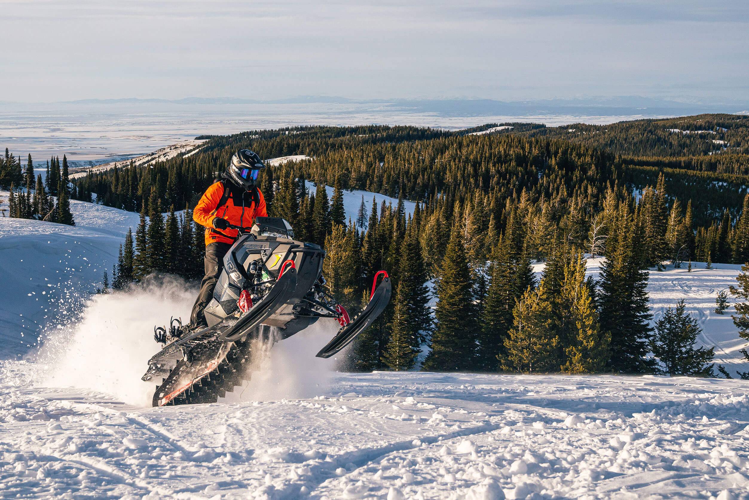 a man in a bright orange snow jacket, gets air on a snowmobile and kicks up snow with green pine trees in the background