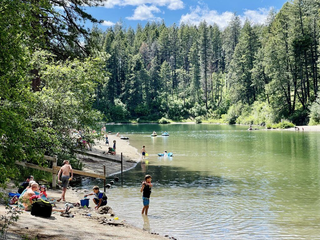 Several families in swimsuits sit and stand on the shore of Lake Pend Oreille, playing in the water and sun, at Farragut State Park. 