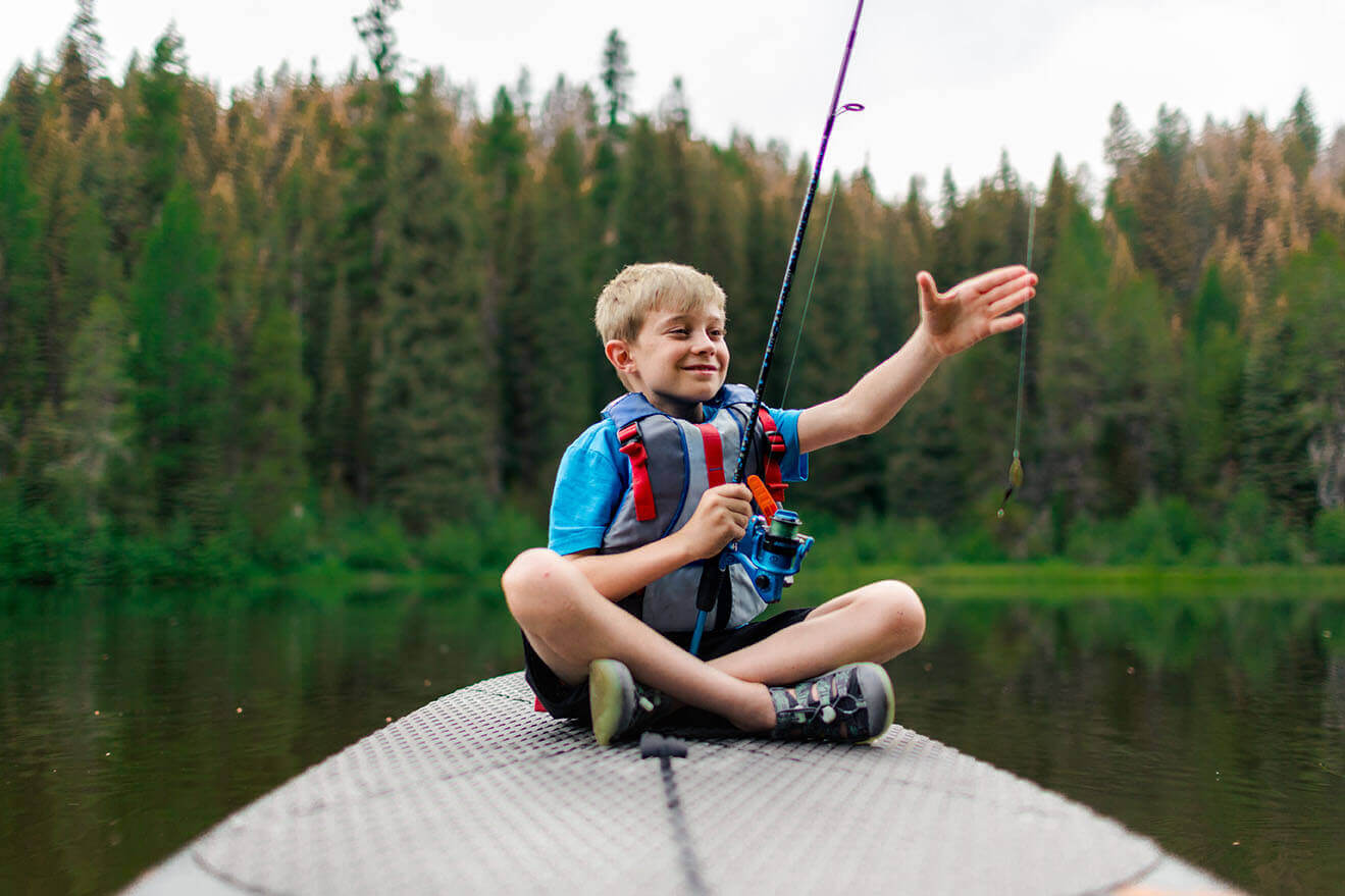 A boy sits on a paddleboard, smiling as he holds a fishing line at the Tripod Reservoir.