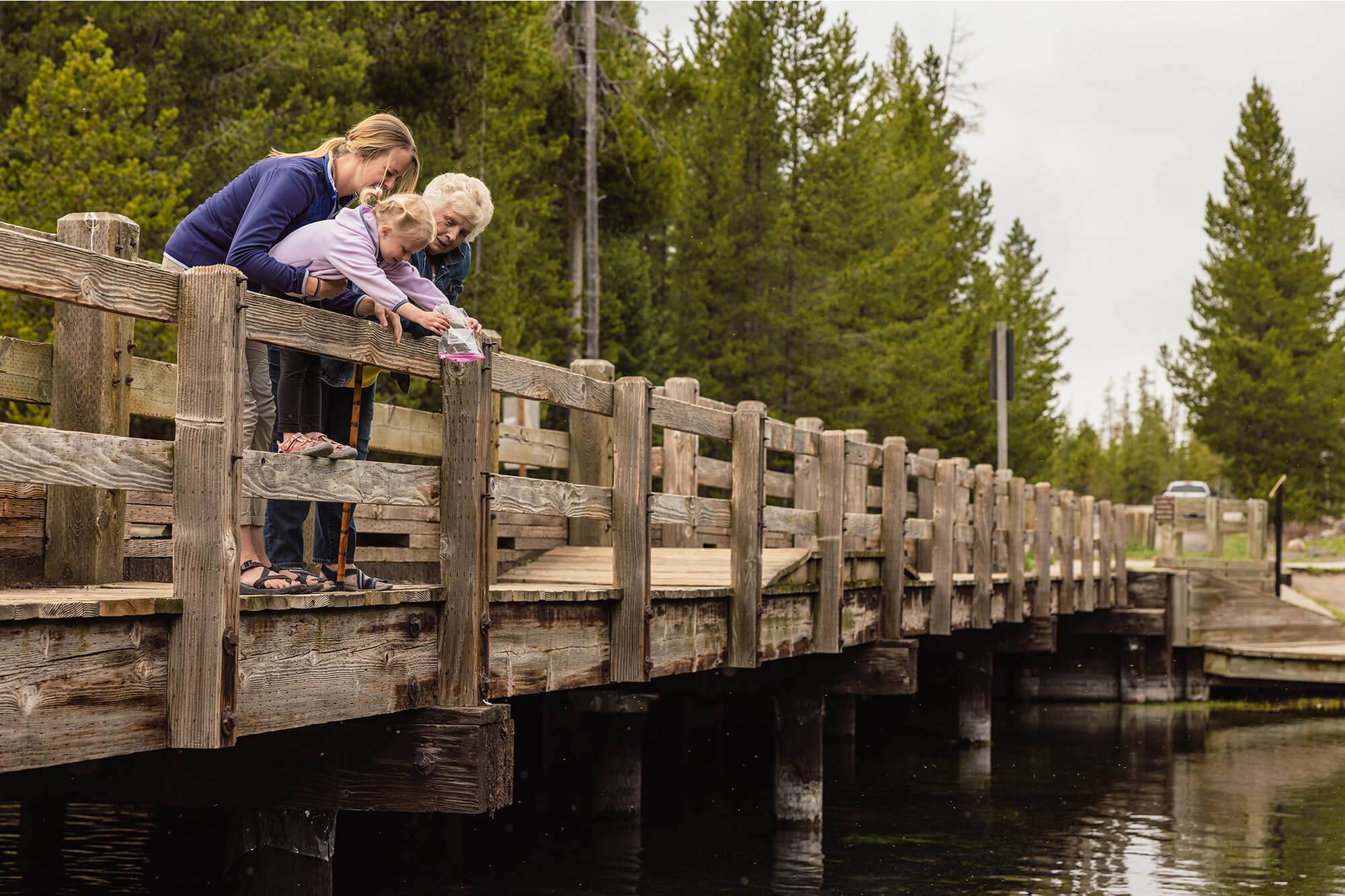 Three generations of women stand on a wooden bridge and feed the fish below.