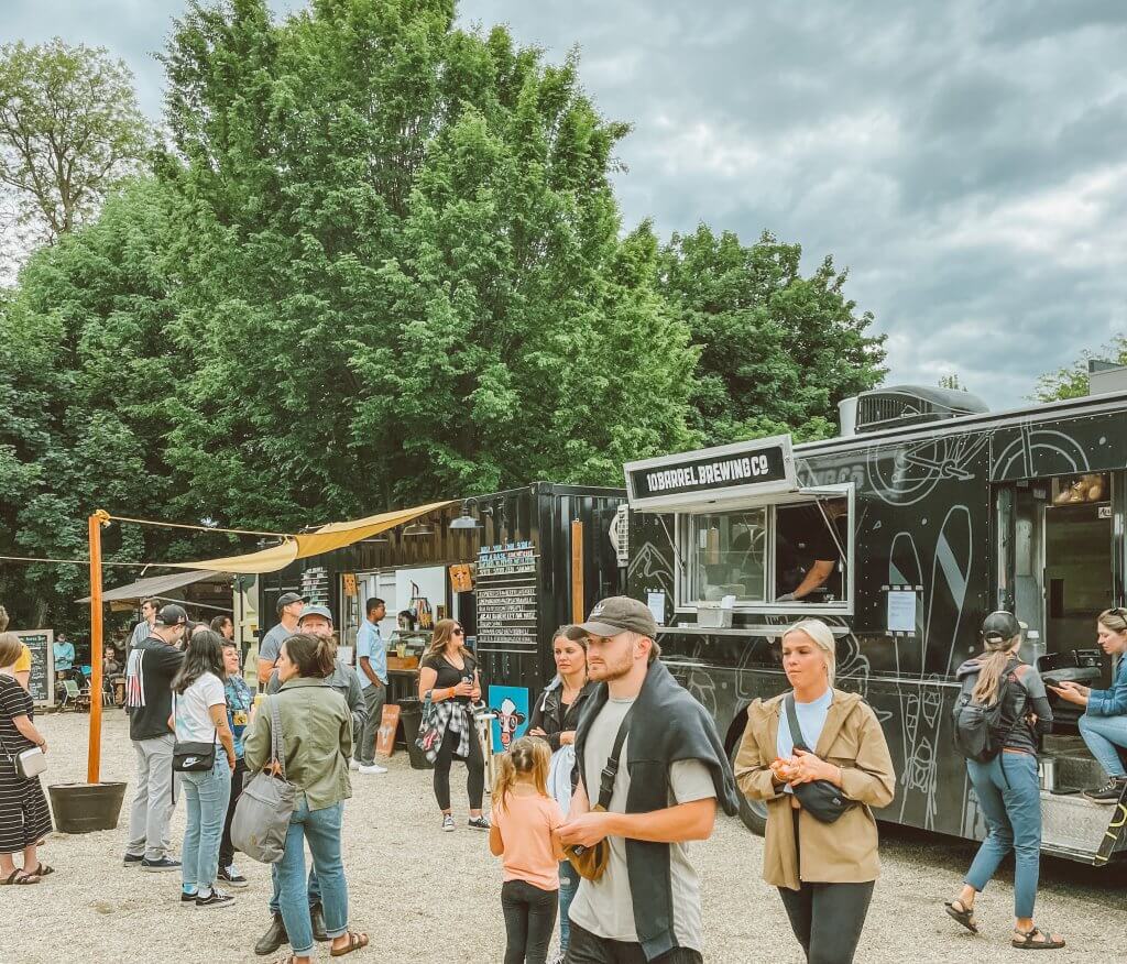 People mingle at the Green Acres Food Truck Park with a black food truck in the background.
