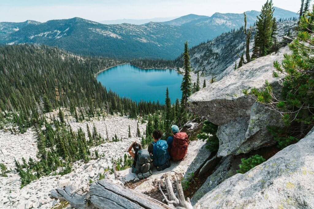 three people with backpacking gear on sitting on a rock looking at Harrison Lake far off in the distance