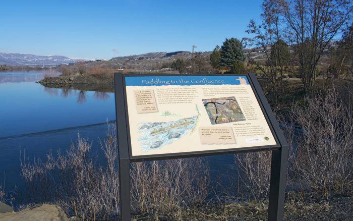 An interpretive trail sign along a body of water surrounded by trees and bare bushes.