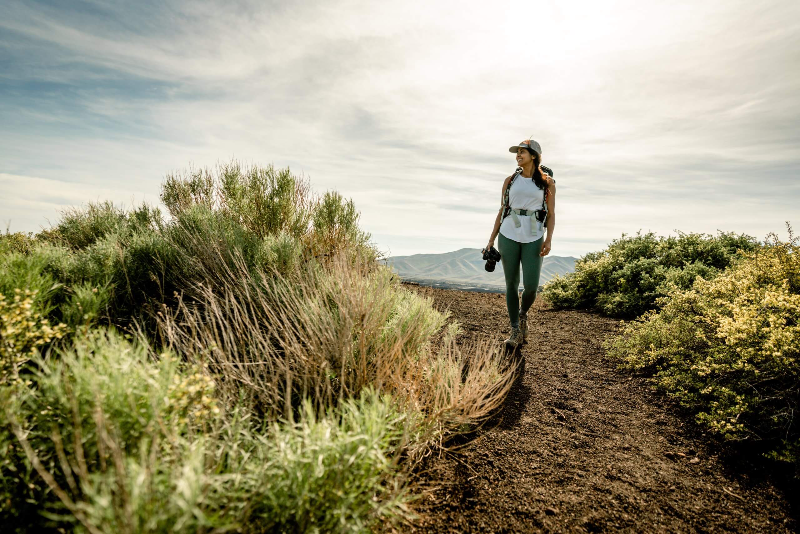 A woman hiking alone on a trail between green brush under sunny skies.