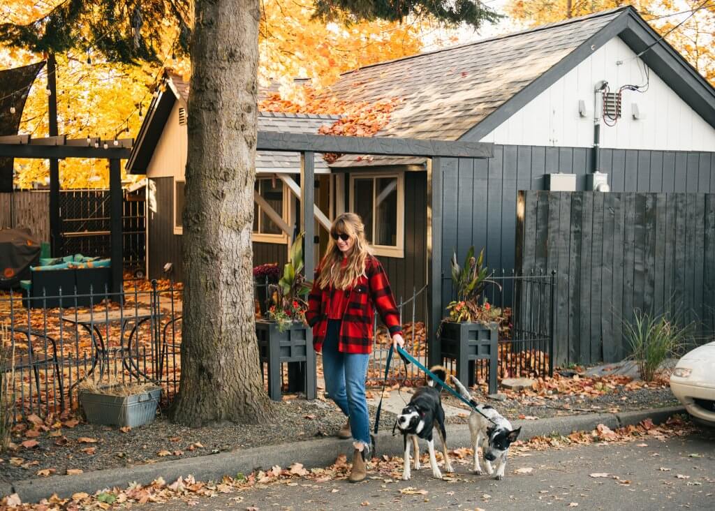 woman in red and black checked flannel walking two dogs on leashes on city street dusted with leaves