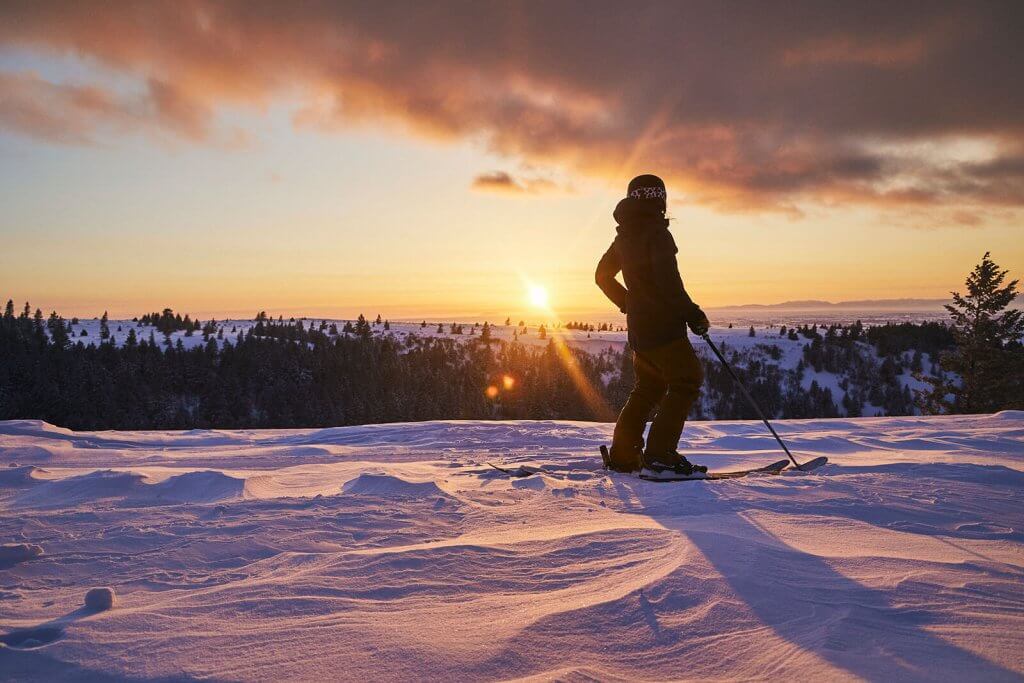 A person in ski gear looks out over a snow-blanketed landscape during sunrise.
