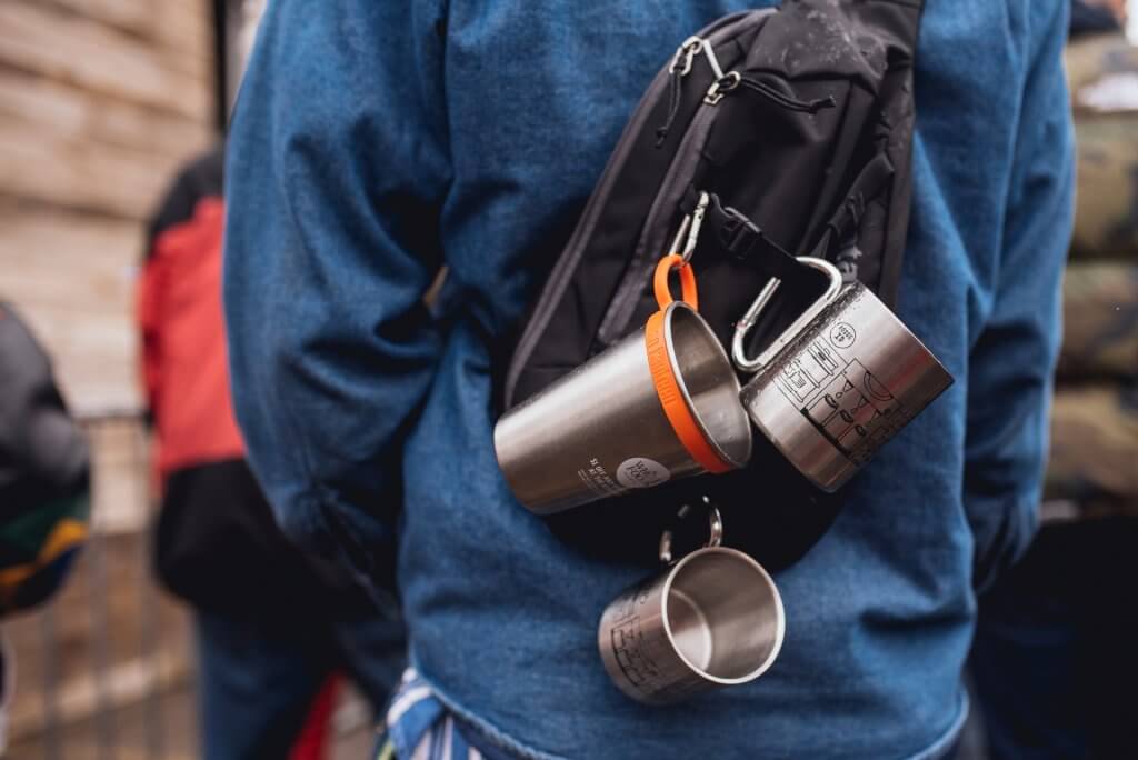 Two steel mugs and one steel cup clipped to a black backpack. A person in a blue jacket is wearing the backpack.