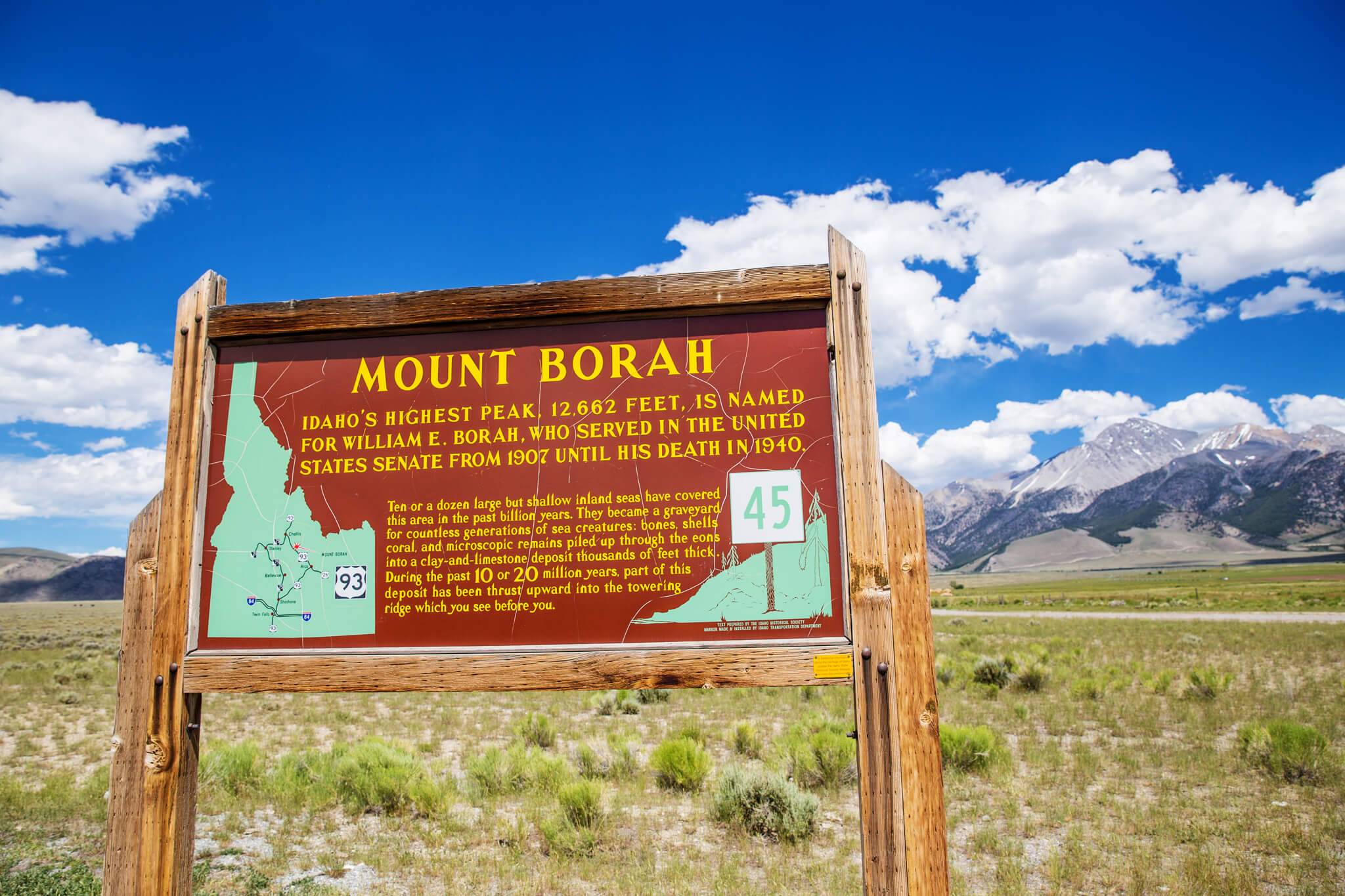 A wooden sign sharing facts and a map of Mount Borah, with Mount Borah during a clear, blue day in the background.