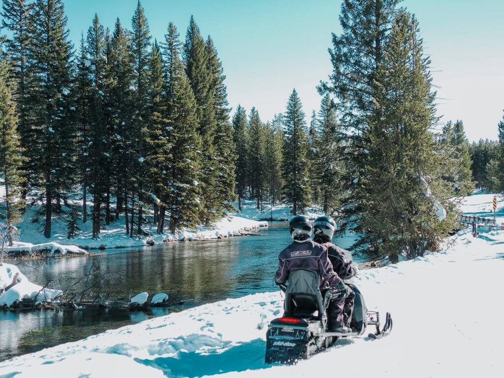 two people in winter gear sitting on snowmobile on snowy path looking at river