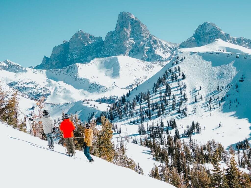three skiers standing in a line on a snowy ski run at grand targhee looking at the teton mountains