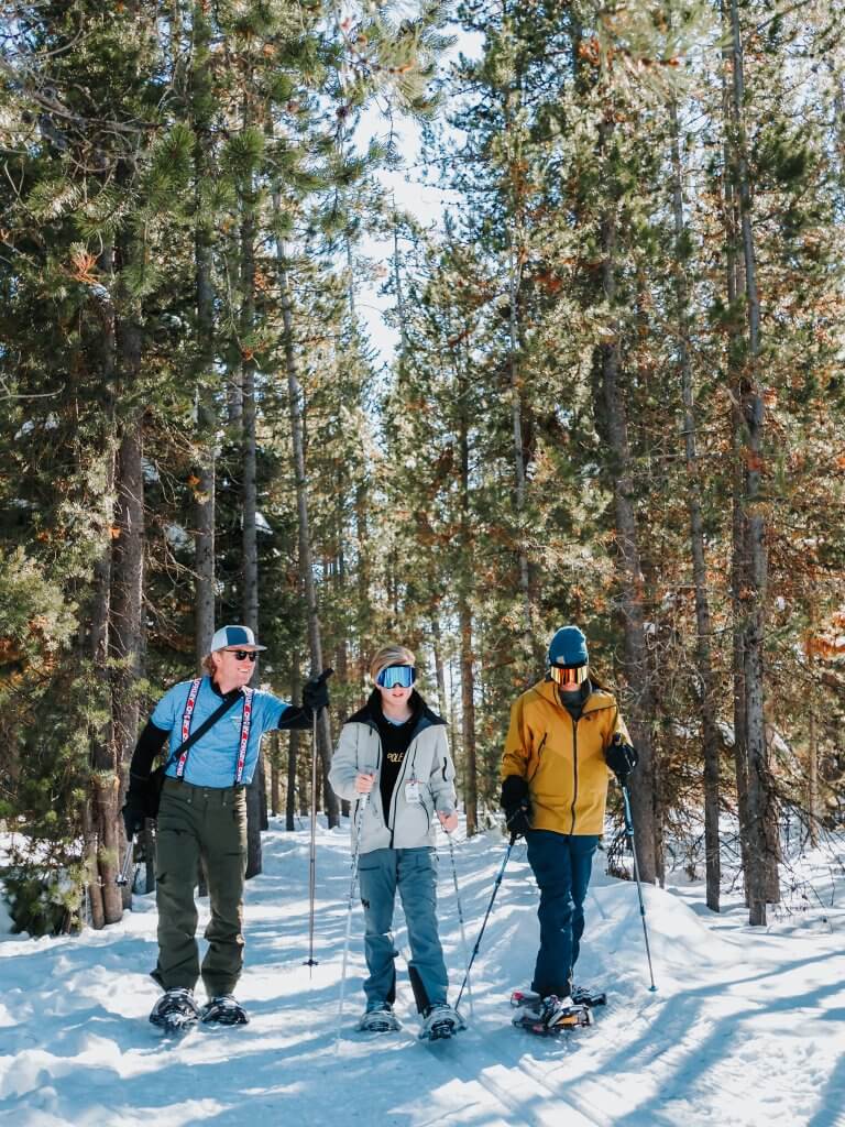 three people snowshoeing in snowy forest with bright blue sky in background