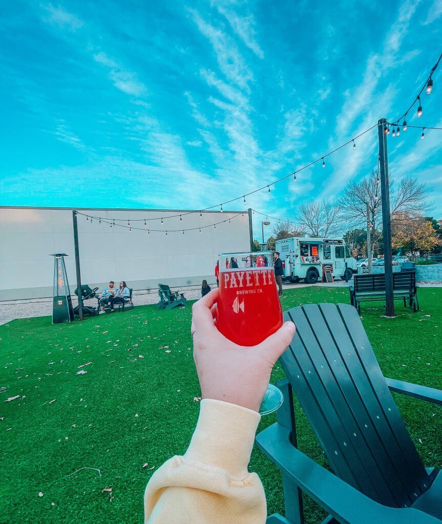 A woman's hand holds of goblet of reddish orange beer in the air with a food truck and blue sky in the background.