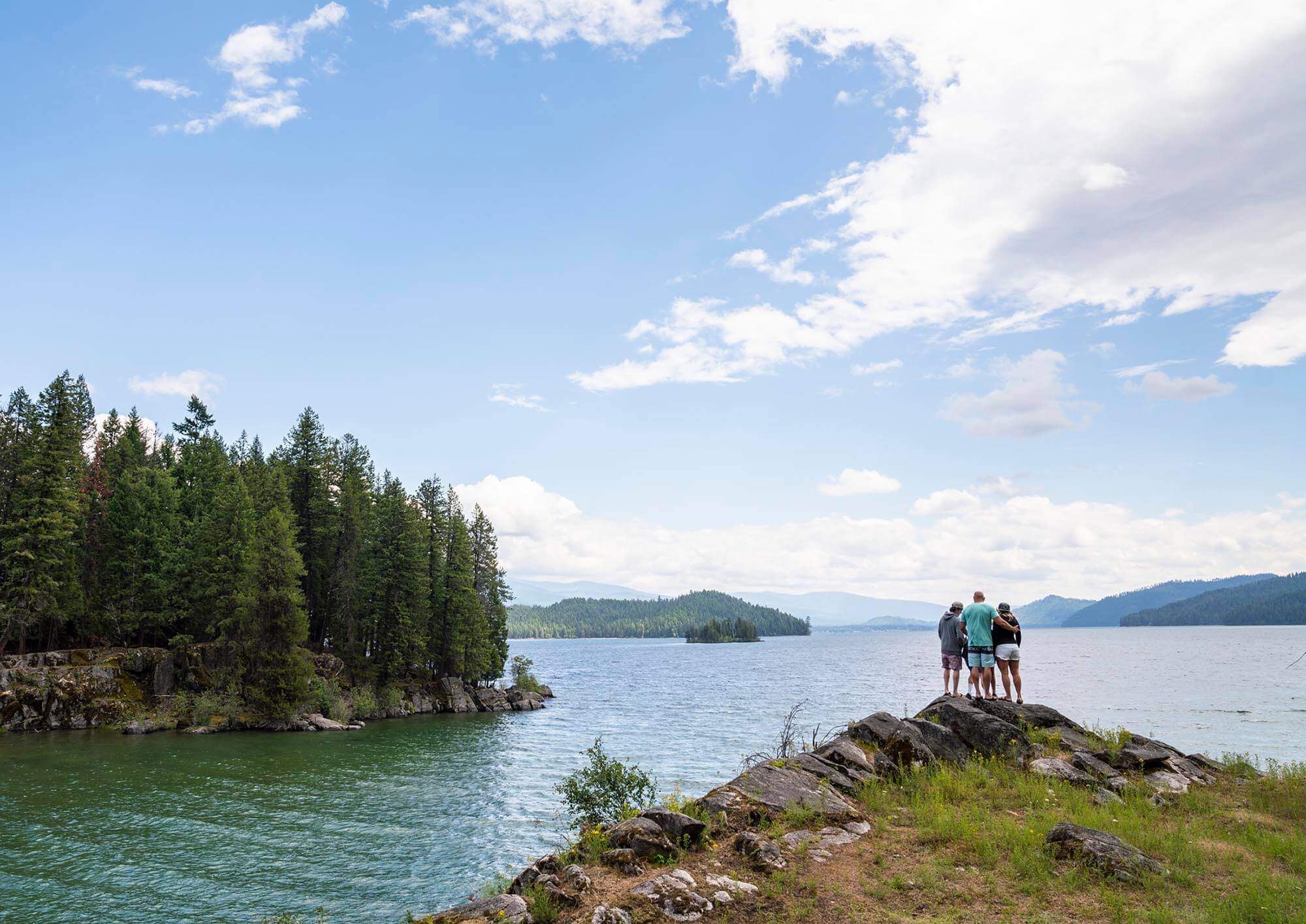 Four people stand in an embrace overlooking a scenic lake with small, forested islands.