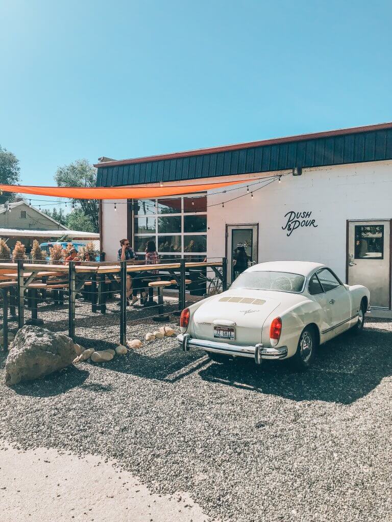 Exterior of Push and Pour coffee shop with white stucco exterior wall, an orange canopy covering picnic tables all next to a vintage white car.