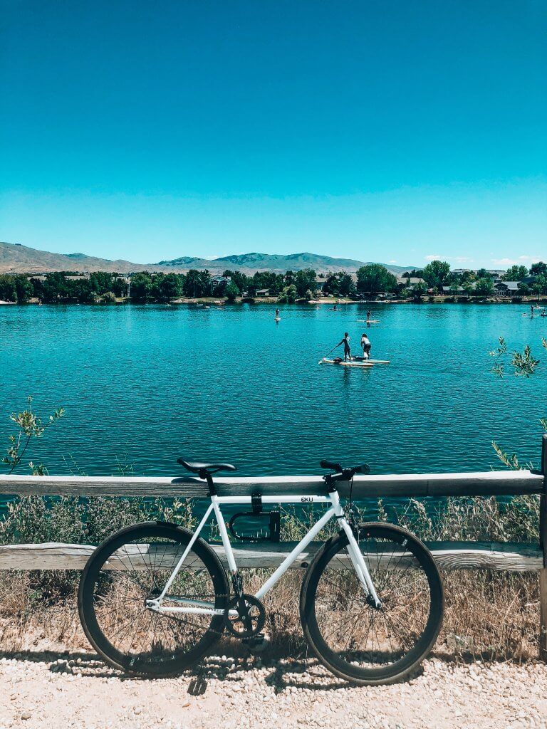 A white bicycle leans against a wood fence while a very large pond sits in the background with paddleboarders.