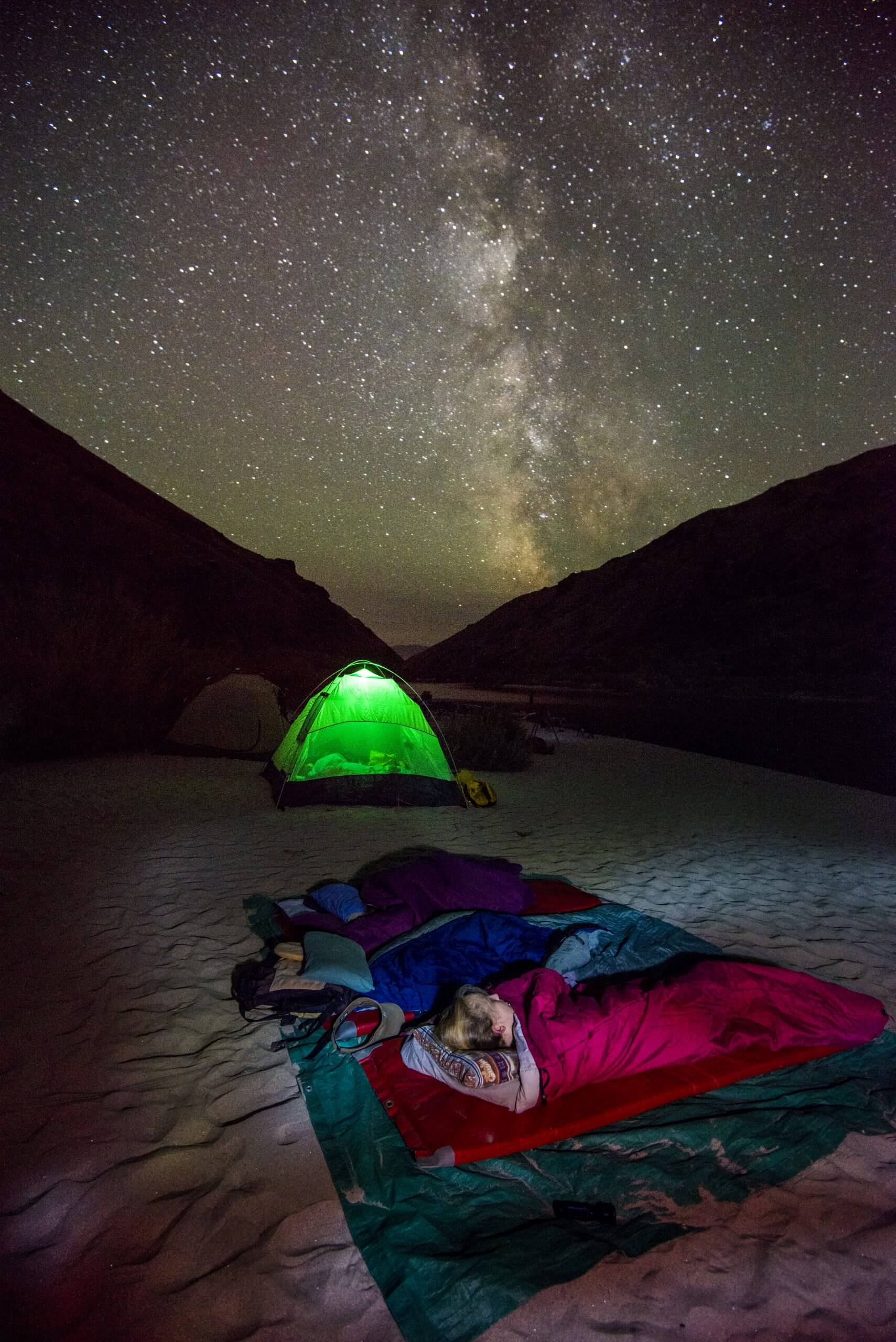 people sleep under the stars in sleeping bags with an illuminated green tent in the background