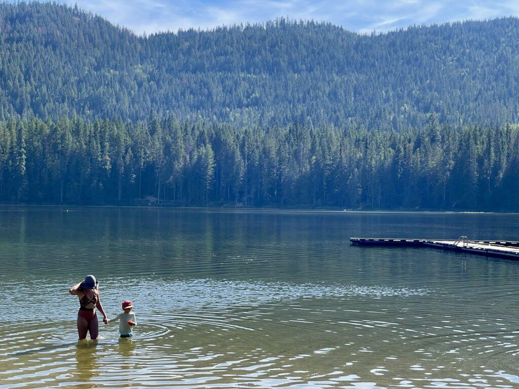 A mom and her son stand in the lake at Round Lake State Park with a dock floating in the water to the right. 