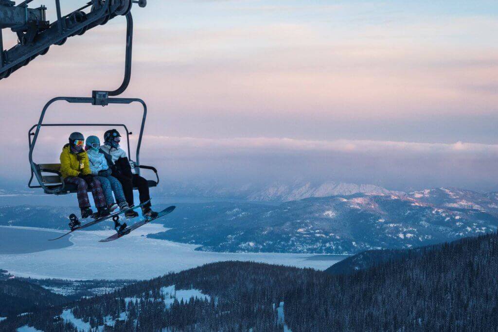 Three people in ski gear sitting on a chair lift during sunrise with mountains and a lake behind them.