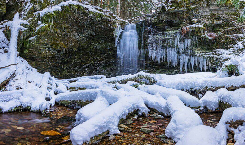 A view of the frozen waters of Shadow Falls with snow covered rocks and icicles in the foreground.