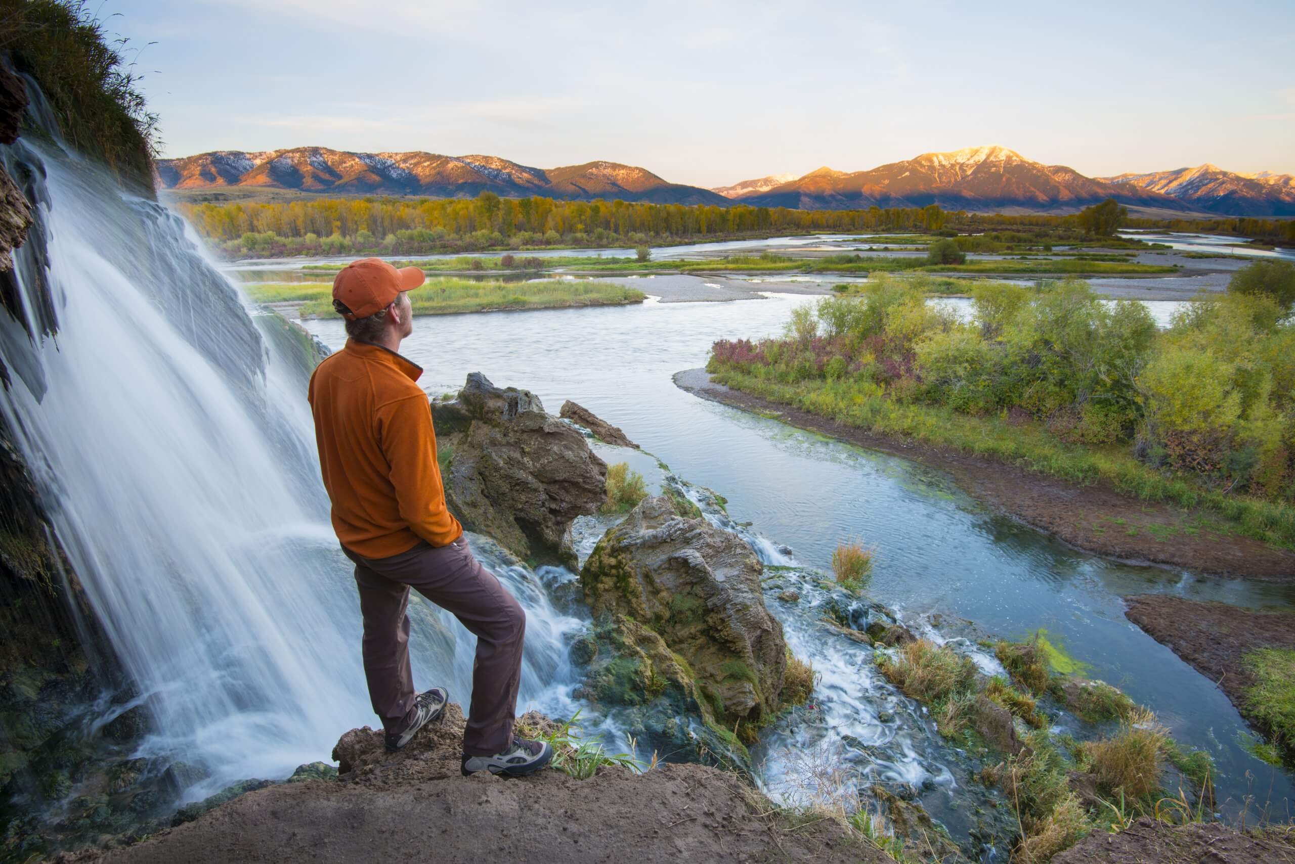 A man in an orange jacket and orange baseball hat stands next to a waterfall, overlooking a valley