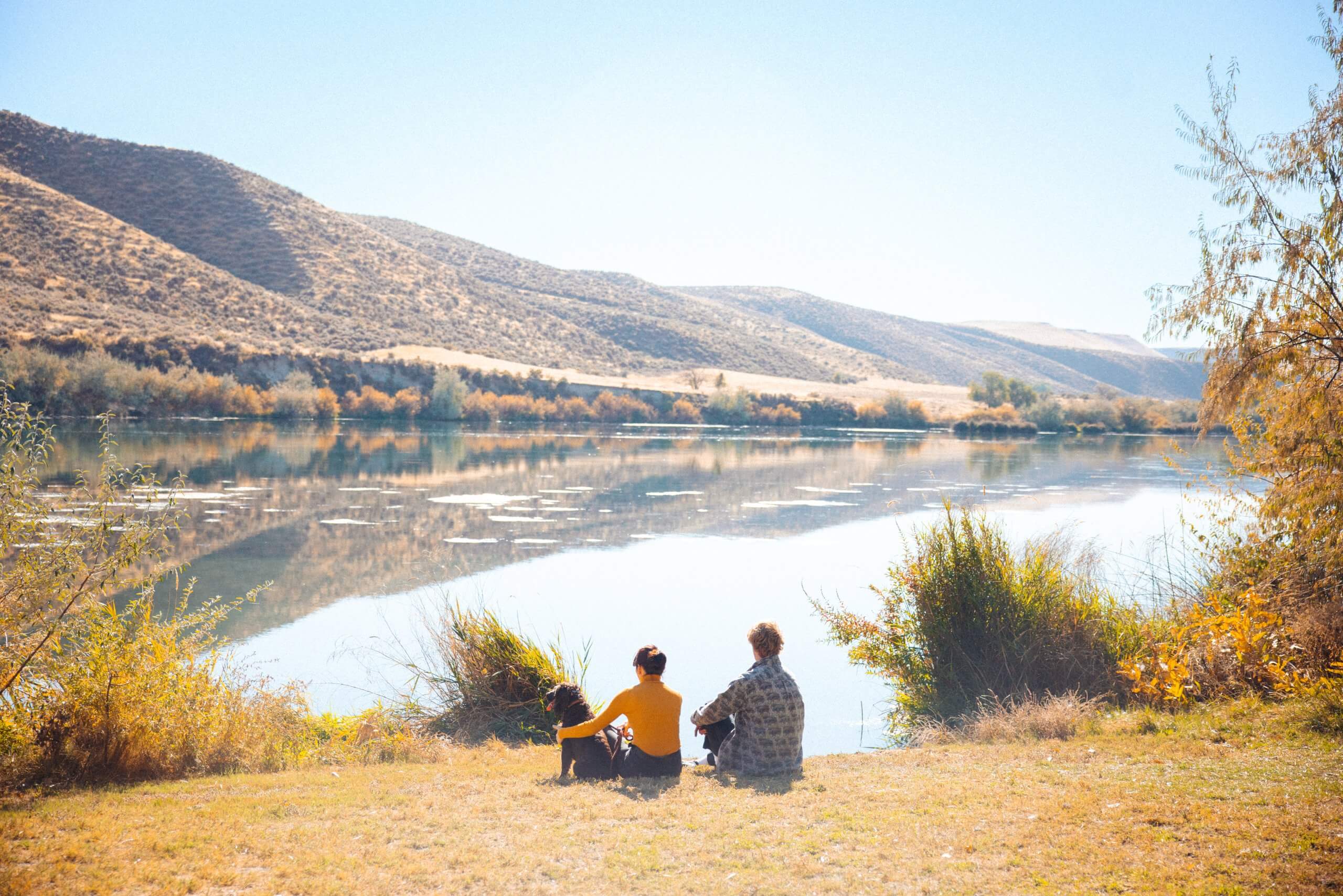 Two people sit with a dog and look out at a body of water in the fall.