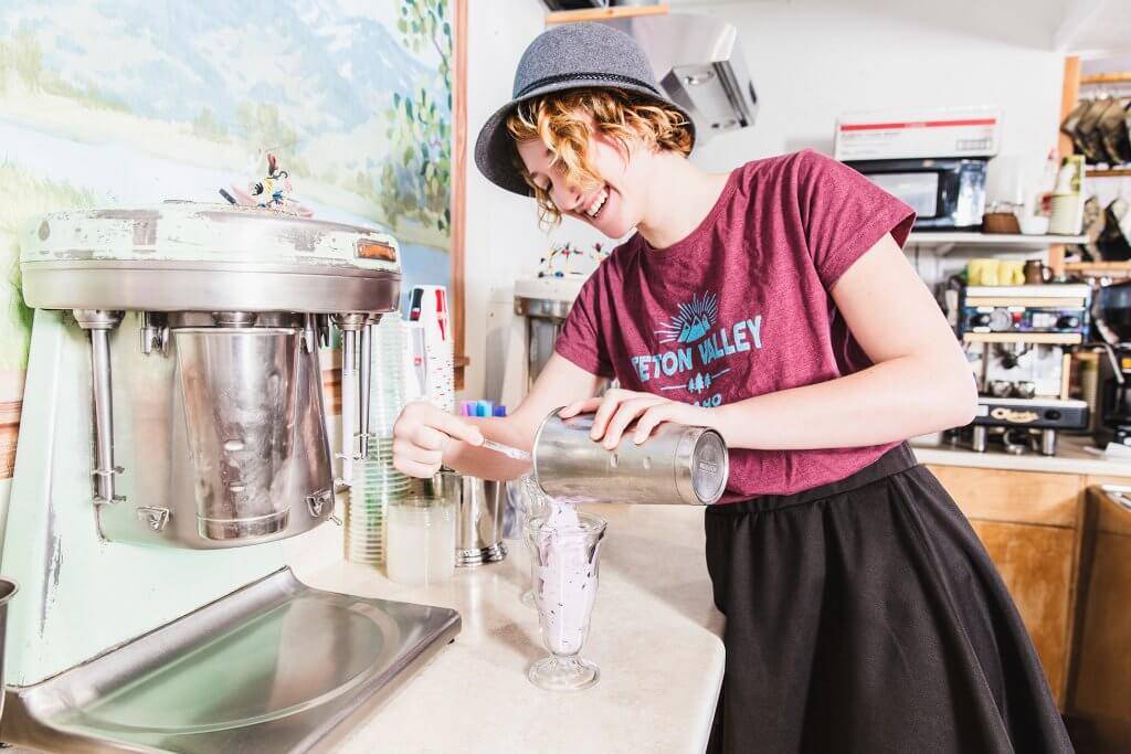 a woman in a maroon Teton Valley shirt, smiles and pours a milkshake into a glass