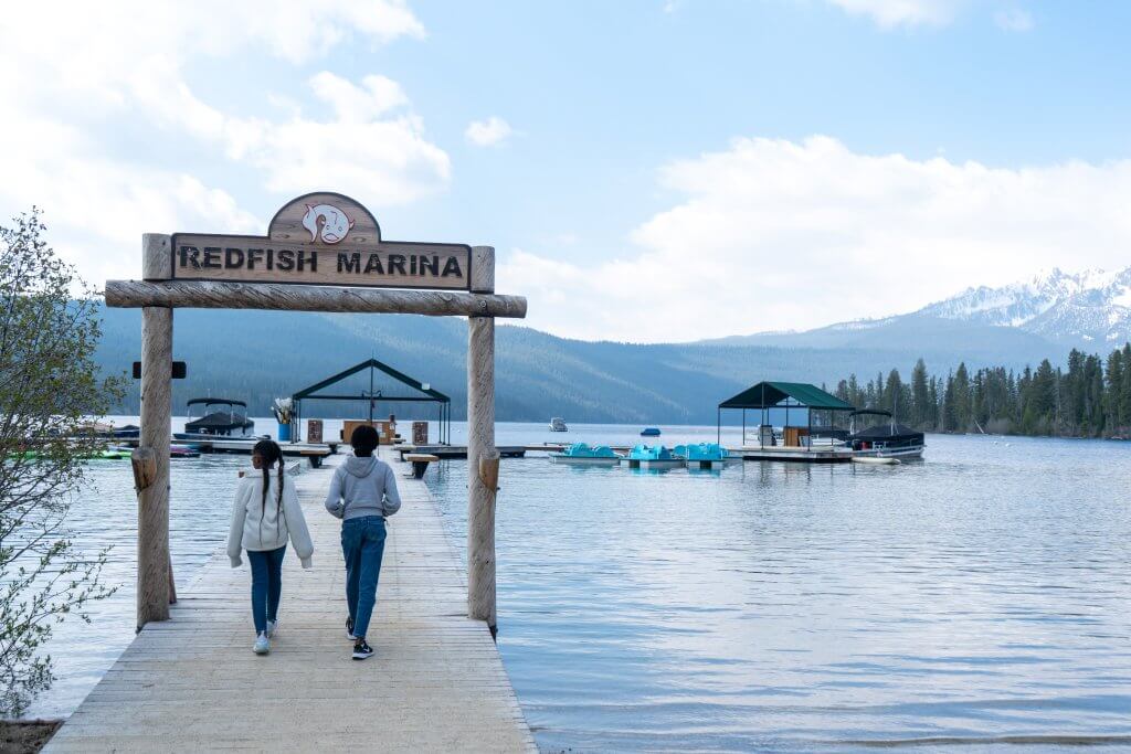 two young girls in jeans and sweatpants walking on wooden boardwalk under a sign that says "redfish marina" with the lake and Sawtooth mountains in the distance