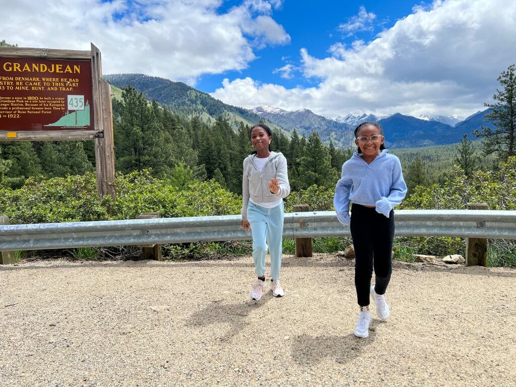 Two young girls are walking toward the camera. Rugged mountains, lush forest and an informational sign are behind them at a scenic overlook.