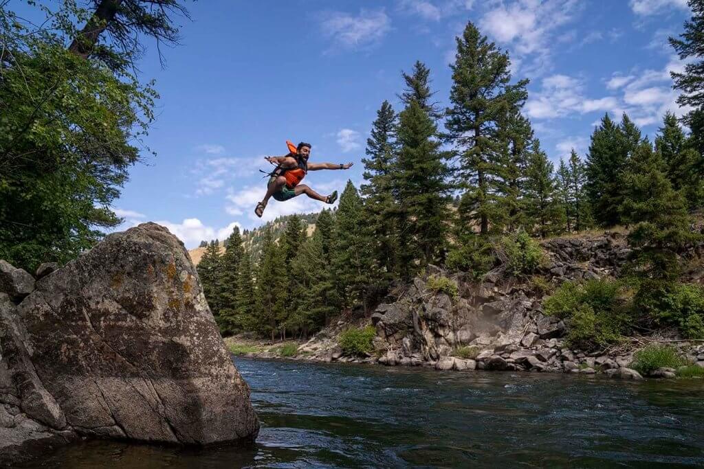 A man wearing a life jacket jumping into a body of water surrounded by large rocks and tall trees.