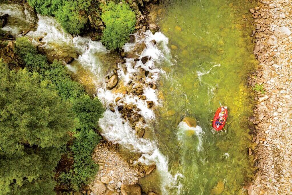 a birds-eye view of people paddling an orange raft on whitewater river lined with trees